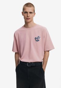 Pink cotton t-shirt with a crew neck and short sleeves, featuring a black and red graphic heart design on the left chest. Worn with dark pants.