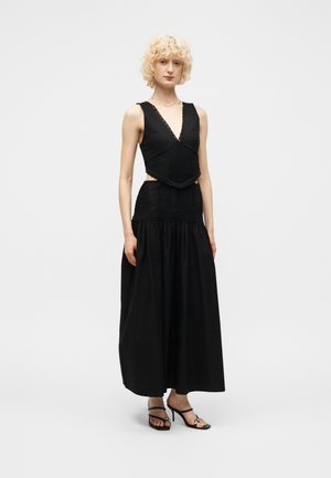 Göndör szőke hajú nő ujjatlan fekete V-nyakú felsőt és hosszú fekete szoknyát visel magassarkú szandállal, egyszínű háttér előtt áll.