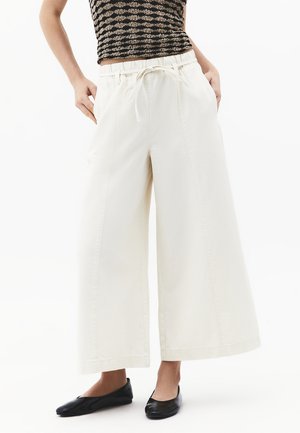 Personne portant un pantalon blanc taille haute à jambes larges avec cordon de serrage et des ballerines noires, debout avec les mains dans les poches sur un fond blanc.