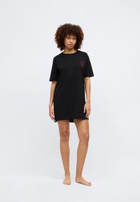 Μαύρο βαμβακερό φόρεμα t-shirt με κοντά μανίκια, έχει κεντημένη μια κόκκινη καρδιά στο αριστερό στήθος και έχει χαλαρή εφαρμογή με πλαινές σχισμές.