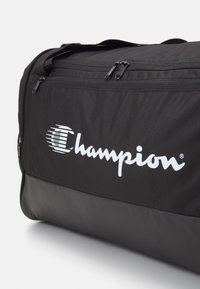 Bolsa de deporte negra con tela texturizada, que presenta un destacado logo blanco de "Champion" y dos aberturas con cremallera negra en la parte superior.
