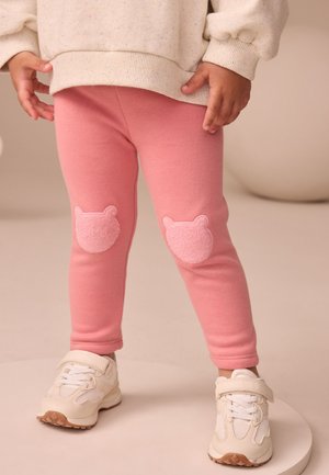 Lyserøde bomuldsleggings med bløde knælapper formet som bamser, kombineret med hvide sneakers med en textureret sål og velcrolukning.