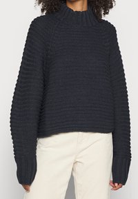 Mörkblå stickad tröja med hög krage, strukturerad ribbning och långa ärmar. Den kortade designen framhäver en avslappnad passform.