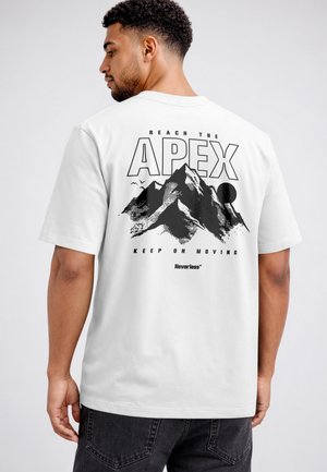 Mann trägt weißes T-Shirt mit schwarzer Berggrafik und dem Text „Reach the Apex, Keep on Moving“ auf dem Rücken, steht vor einfarbigem Hintergrund.