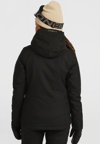 Personne portant une veste d'hiver noire O'Neill avec capuche, un bonnet beige, des lunettes de ski noires et des gants noirs, de dos contre un fond blanc.