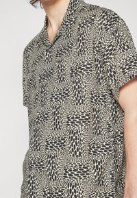 Kratší rukávový košilový top s béžovým a černým potiskem, který zobrazuje abstraktní oválné tvary a patchworkový design.