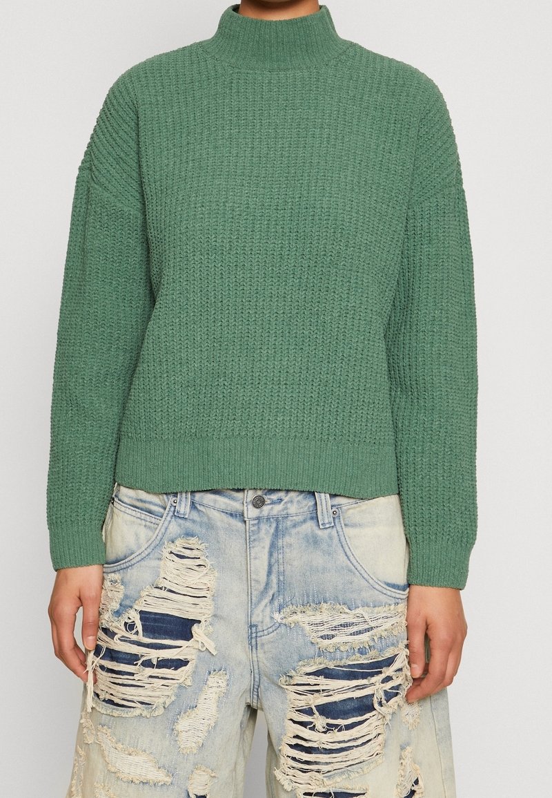 Pull vert en maille avec col montant et détails côtelés, associé à un short en denim délavé clair usé avec bords effilochés et patches.