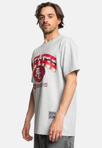 Graues Baumwoll-T-Shirt mit einem auffälligen roten Grafikdesign, das Text und ein rundes Emblem enthält, kurzen Ärmeln und einem Etikett an der Seite.
