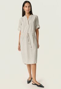 Robe chemise rayée en tissu léger, dotée d'un col montant, de manches courtes retroussées et d'une taille à cordon de serrage. Portée avec des chaussures à talons compensés noires.