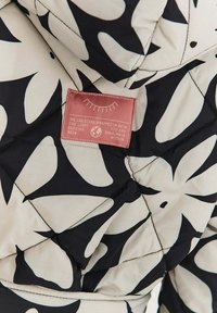 Prošívaná bunda v krémové barvě s velkými černými listovými vzory. Obsahuje růžovou etiketu s textem a stylizovanými grafickými detaily.