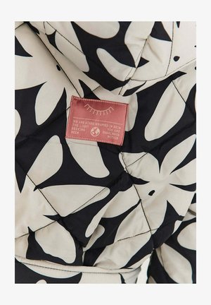 Kurtka pikowana wykonana z kremowego materiału z dużymi czarnymi wzorami liści. Zawiera różową metkę z tekstem i stylizowanymi graficznymi detalami.