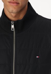 Black padded jacket with a high ribbed collar, silver zipper, and small logo patch featuring red, white, and blue stripes on the chest.