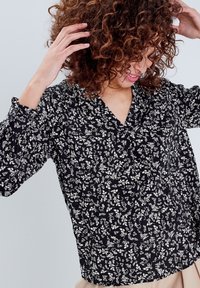 Blouse noire à fleurs avec un col en V et des manches longues bouffantes. Présente de petits motifs floraux blancs et un tissu doux et texturé.