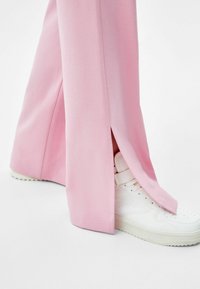 Pink tailored pants with side slits, paired with a white high-top sneaker featuring velcro straps and a textured sole.