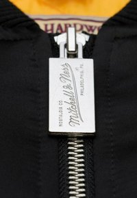 Schwarzer Reißverschluss mit einem silbernen Metall-Zip Pull, der "Mitchell & Ness" eingraviert hat, mit einer strukturierten Oberfläche, der an einem darunterliegenden gelben Stoff befestigt ist.