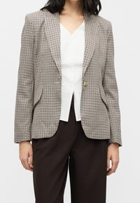 Femme portant un blazer à petits carreaux beige et noir par-dessus un chemisier blanc à col en V et un pantalon marron foncé, debout devant un fond uni.