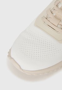 Primer plano de la punta de una zapatilla beige y blanca con tejido perforado y suela de goma texturizada.