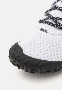 Chaussure de sport blanche avec une tige en mesh, semelle en caoutchouc noir et semelle texturée, lacets avec des motifs contrastants en noir et blanc.