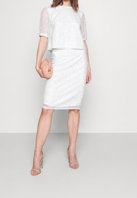 Robe blanche en deux pièces avec un haut court texturé à manches transparentes et une jupe ajustée arrivant au genou. La tenue est accompagnée d'une pochette rose clair.