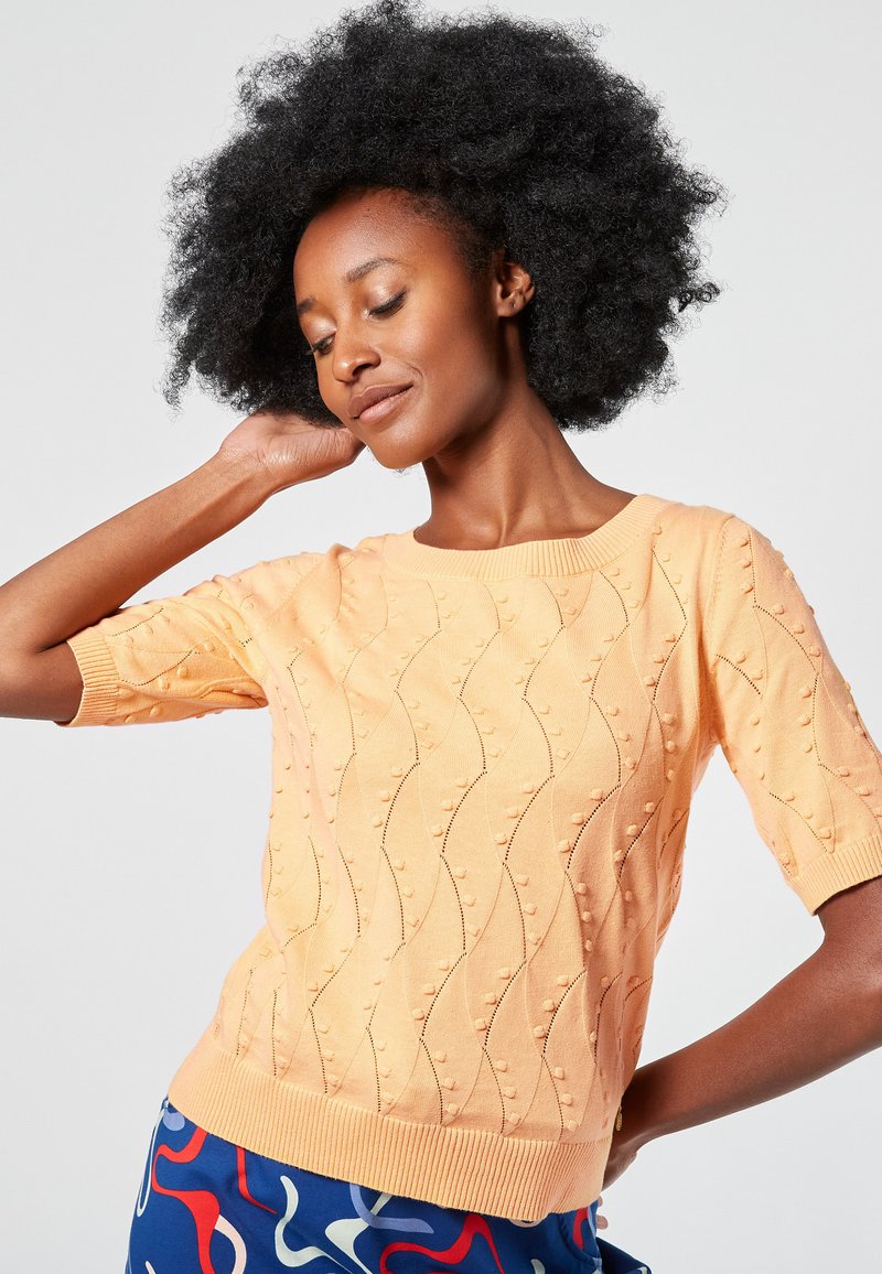 Lyhythihainen persikanvärinen neulepaita, jossa on aaltoilukuosi kohotetuilla pisteillä, ribbaus kaula-aukossa ja helmassa. Yhdistetty värikkäisiin abstraktin kuvioisiin housuihin.