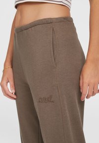 Pruunid dressipüksid, millel on nööriga talje, küljetaskud ja broderi logo detailid. Pehme kangas, millel on sile tekstuur.