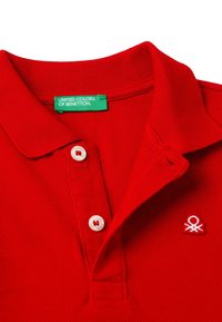 Červená polo tričko s texturovanou tkaninou, dvěma bílými knoflíky a malým bílým výšivkovým logem na levé straně hrudi. Zelená značka uvnitř límce.