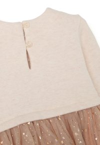 Helles beige Oberteil mit einem runden Ausschnitt, hinten mit zwei Knöpfen versehen; layered mit einem Tüllrock, der mit goldenen Glitzerpartikeln verziert ist.