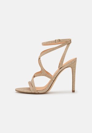 Glitrende gylden højhælet sandal med krydsede remme, åben tå og ankrem. Glat sål og struktureret hæl, elegant design.