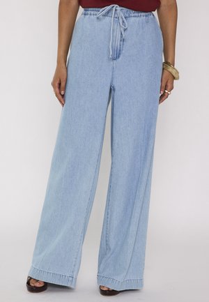 Femme portant un jean bleu clair à jambes larges avec taille élastique et cordon, assorti à des chaussures ouvertes foncées et un bracelet au poignet.