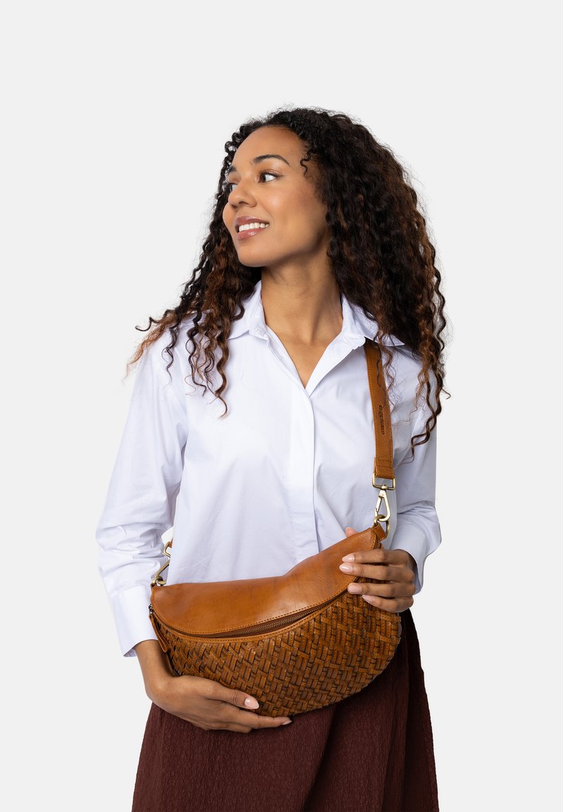 Borsa a spalla in pelle marrone chiaro con texture intrecciata, forma curvata, chiusura con zip e tracolla regolabile. Il modello indossa una camicia bianca e una gonna marrone.