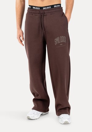 Mężczyzna noszący brązowe, luźne spodnie dresowe z logo "SMILODOX" na udzie i czarnym elastycznym pasem, zestawione z białymi sneakersami.