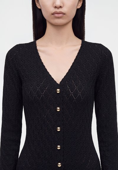 Femme portant un cardigan en maille noire texturée avec un profond décolleté en V et cinq boutons dorés brillants sur le devant.