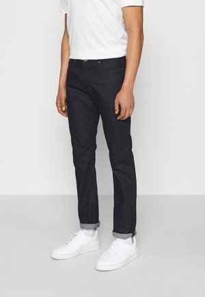 Muž v černých ohrnutých džínách, bílých teniskách a bílém tričku stojí před jednoduchým světlým pozadím.