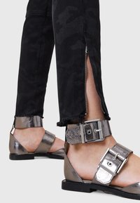 Jeans skinny de camuflaje negro con cremalleras plateadas en los laterales, combinados con sandalias metálicas plateadas que tienen tiras anchas y detalles de hebilla.