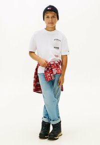 T-shirt bianco a maniche corte con toppe logo, jeans azzurri, camicia a quadri rossa legata in vita, stivali neri e berretto blu navy.