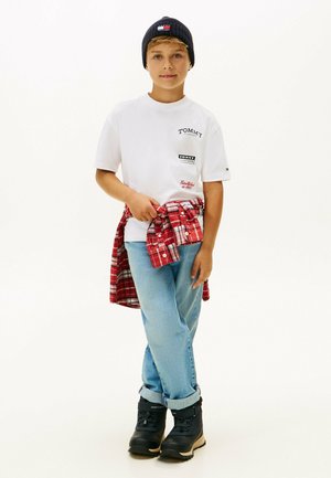 Hvid t-shirt med korte ærmer og logo patches, lyseblå jeans, rødternet skjorte bundet om taljen, sorte støvler og marineblå hue.