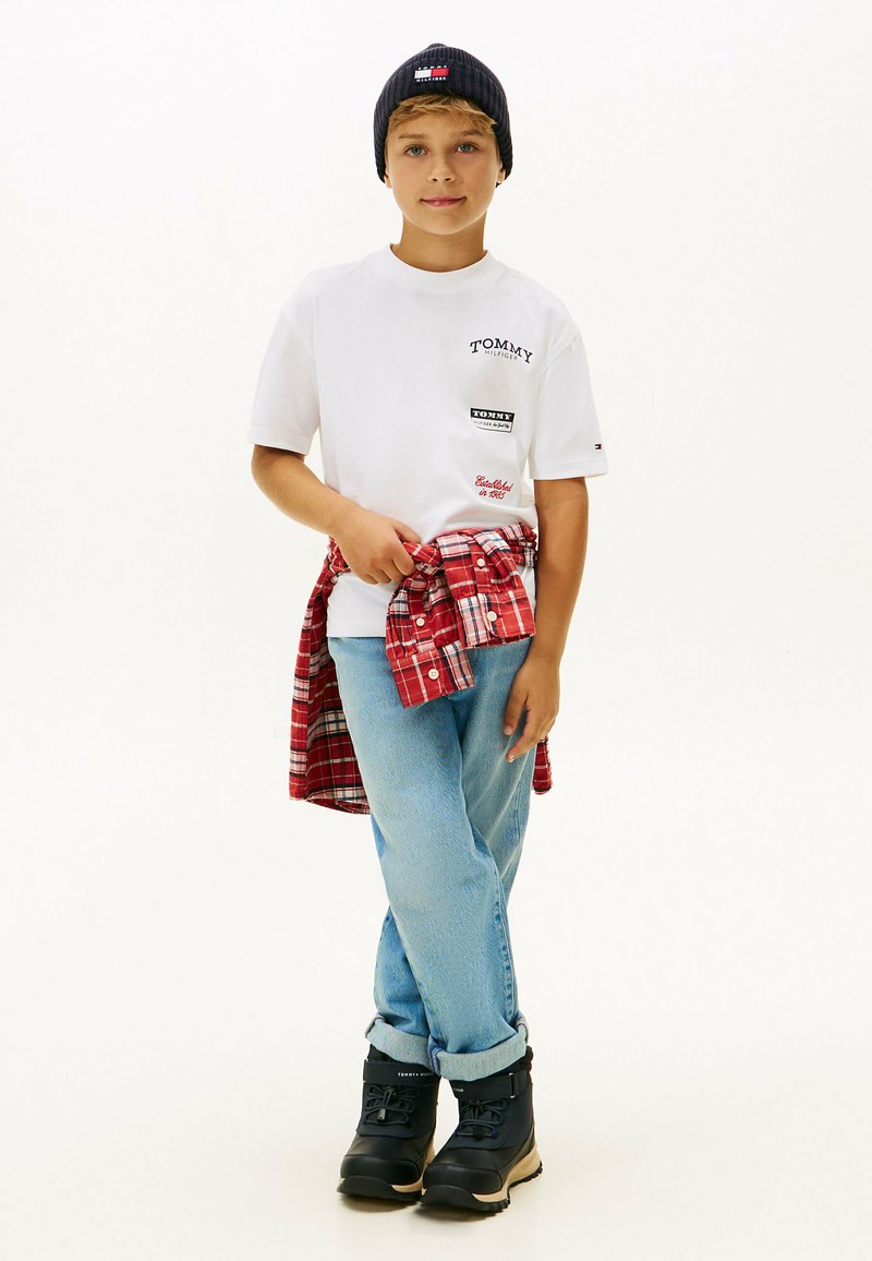 T-shirt bianco a maniche corte con toppe logo, jeans azzurri, camicia a quadri rossa legata in vita, stivali neri e berretto blu navy.