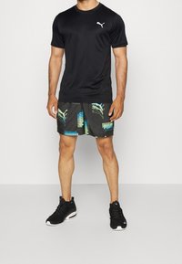 Camiseta deportiva negra de manga corta, combinada con pantalones cortos negros que presentan logotipos de Puma en azul y verde y un patrón geométrico. Zapatillas negras.