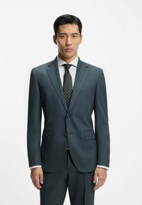 Mann in einem dunkelgrünen, maßgeschneiderten Anzug mit einer schwarzen gemusterten Krawatte und weißem Hemd, stehend vor einem schlichten weißen Hintergrund.