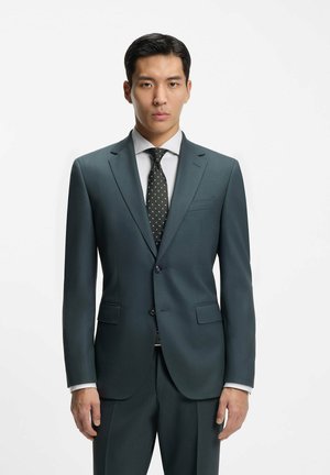 Man in donkergroen op maat gemaakt pak met zwarte patroonstropdas en wit overhemd, staand tegen een effen witte achtergrond.