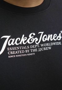 Mustern kõrge kaelusega must sviiter, millel on valge trükitud tekst: "Jack & Jones ESSENTIALS DEPT. WORLDWIDE" ja "SINCE NINETEEN NINETY."