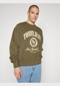 Felpa verde oliva con polsini e orlo a costine, con un grande logo giallo centrato che legge "TOMMY JEANS" e "New York".