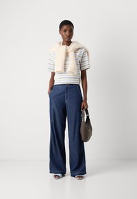 Gestreiftes kurzärmliges Hemd in Blau und Weiß, mit einem cremefarbenen Schal drapiert, kombiniert mit weit geschnittenen, dunklen Denim-Hosen und blauen Absatzsandalen. Braune Handtasche in der Hand.