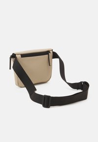 Sac bandoulière beige avec fermeture éclair, sangle noire réglable, texture lisse et design minimaliste. Dispose d'une poche avant.