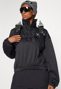 Pullover nero a collo alto con polsini elastici, abbinato a un berretto grigio. Presenta due borracce d'acqua attaccate a un'imbracatura.