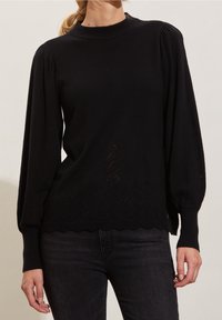 Pull en tricot noir avec des manches longues bouffantes, un col haut, un motif texturé le long de l'ourlet et un bord inférieur à volants.