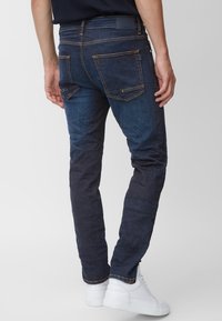 Los jeans de denim azul oscuro presentan un ajuste slim, dos bolsillos traseros y costuras de contraste. Se llevan con zapatillas blancas, mostrando un estilo casual.