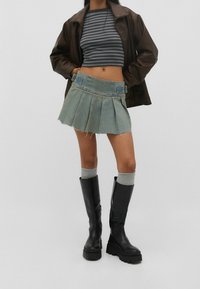 Personne portant un haut rayé court, une veste en cuir marron, une mini-jupe en jean plissée, des chaussettes grises et des bottes noires chunky montantes jusqu'aux genoux.