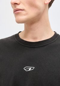 Κοντινό πλάνο του κάτω μέρους του προσώπου και του λαιμού ενός ατόμου που φοράει μαύρο μπλουζάκι με στρογγυλή λαιμόκοψη και μικρό λευκό γεωμετρικό λογότυπο στο στήθος.