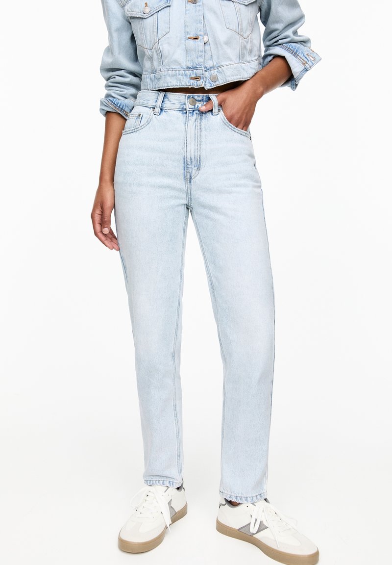 Personne portant un jean taille haute bleu clair, une veste courte en denim, des baskets blanches, debout avec une main dans la poche sur fond blanc.